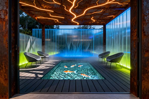 Covered patio framed by weathered corten steel, its canopy etched with branching orange LED lines. Frosted, ribbed polycarbonate walls edge‑lit in electric blue and acid lime encircle a rectangular sunken koi pond lined with teal glass mosaic, set into charcoal composite decking and flanked symmetrically by four dark woven lounge chairs, with low drifting mist pooling at the base of the walls and trees faintly visible beyond.