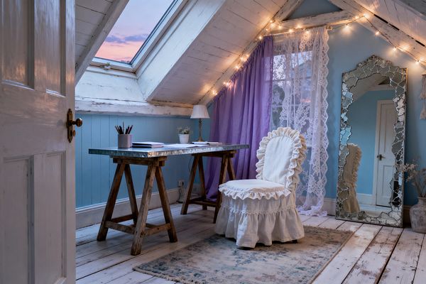 A small attic home office features whitewashed beams and a skylight above a rustic desk with weathered wooden trestle legs and a galvanized metal top. A white ruffled slipcovered chair faces the desk. Sky-blue walls meet bleached plank flooring with a faded blue rug. Lavender curtains and lace drapes hang by the window, with warm string lights along the rafters. A tall distressed mirror leans nearby, plus a brass lamp and a small potted plant.