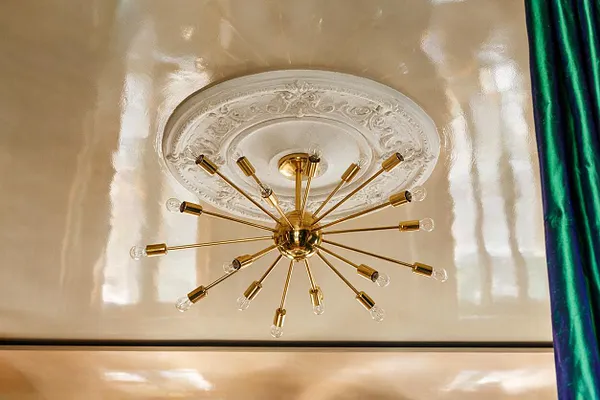 A gold starburst ceiling light with multiple slim arms and exposed bulbs is mounted beneath an ornate white plaster medallion. The glossy cream ceiling reflects light, and a rich green velvet curtain appears along the right edge.