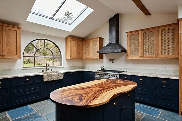 A sunlit transitional kitchen with deep navy Shaker base cabinets and brass pulls, topped with light stone countertops. Upper cabinets in natural oak feature cane panels. A rough-hewn stone farmhouse sink sits beneath an arched black-steel window, with a large skylight above. A blackened steel range hood, pro-style range, and brass pot filler line one wall. The island has a sculptural live-edge wood top, and the floor is blue-gray slate tile.