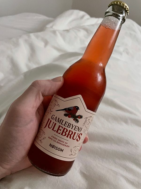 A hand holds a bottle of Gamlebyens Julebrus soda against a soft, white bedspread.