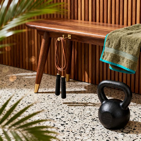 Close-up of a mid-century home gym corner featuring a walnut bench with tapered legs and brass-capped feet in front of a vertical walnut slat wall. A small brass hook beneath the seat holds a leather jump rope, while an olive terry towel with teal trim is draped over the bench. On a light speckled terrazzo floor, a matte black cast-iron kettlebell sits nearby as sunlight and blurred plant leaves cast soft shadows.