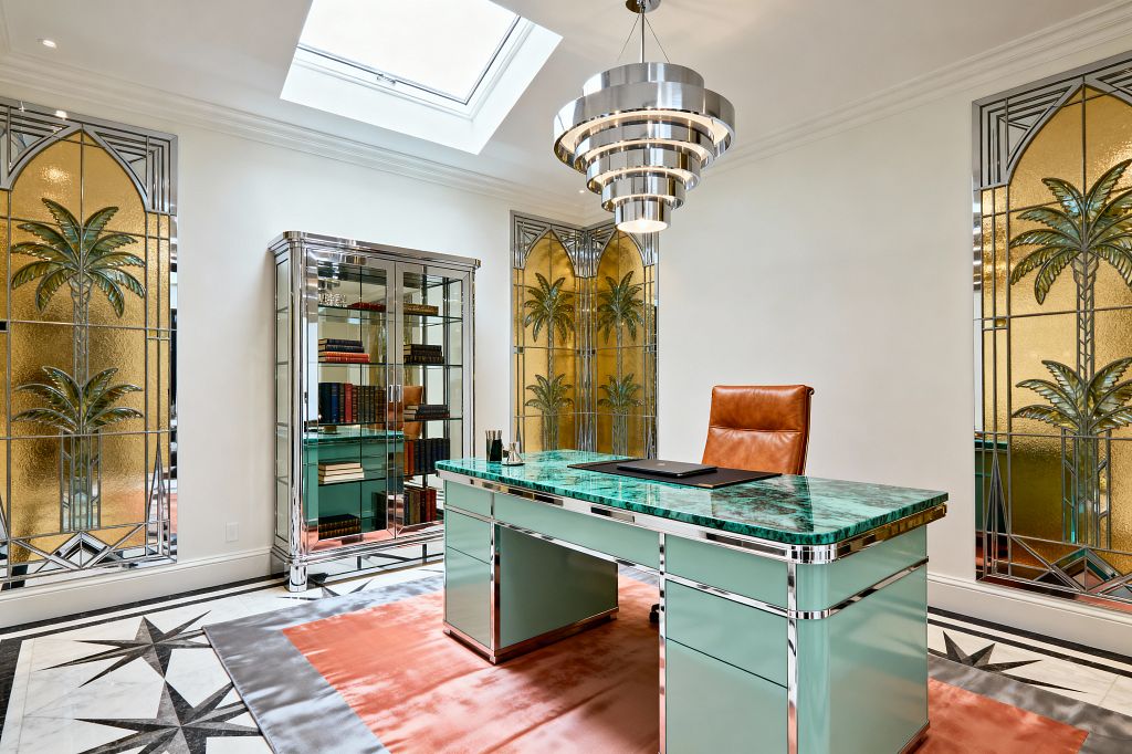 A bright Art Deco home office centers on a mint lacquer desk with a malachite-patterned stone top and chrome trim, paired with a cognac leather chair. A tiered chrome chandelier hangs beneath a skylight, reflecting light onto a coral rug and white marble floor with black starburst inlays. A mirrored glass cabinet displays books on the left. Amber stained-glass panels with stylized palm trees frame the room, adding a glamorous tropical Deco motif.