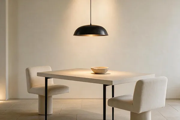 A minimalist dining nook with a rectangular light stone or concrete table, two cream upholstered chairs, and a shallow bowl centered on the tabletop. A black dome pendant light hangs above, casting a warm glow against soft beige walls and large neutral floor tiles.