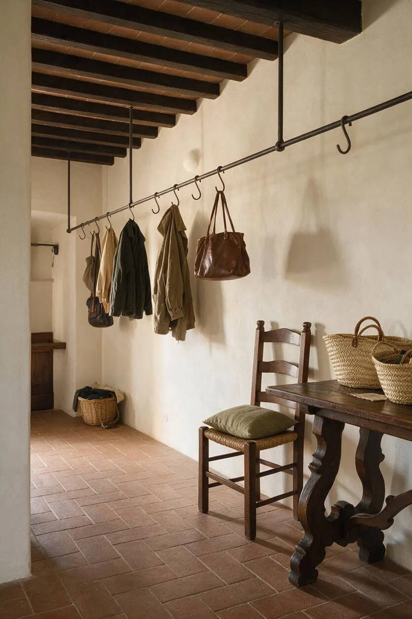 A narrow Mediterranean-style mudroom with terracotta floor tiles, white plaster walls, and dark exposed ceiling beams. A black metal overhead hanging rail holds coats and bags, while a rustic wooden chair, woven baskets, and a heavy wood table add warm, functional character.