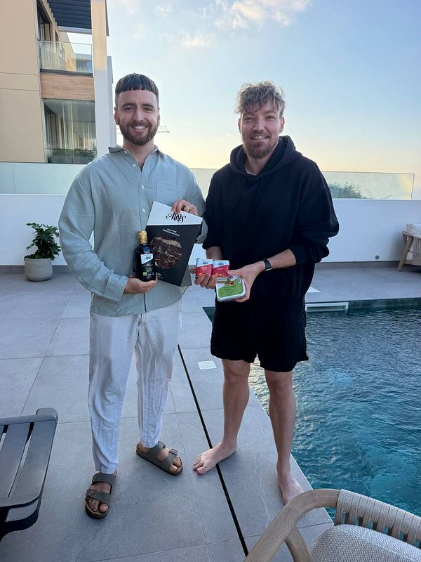 Two men standing by a pool, each holding food and drink items, posing for a photo outdoors.