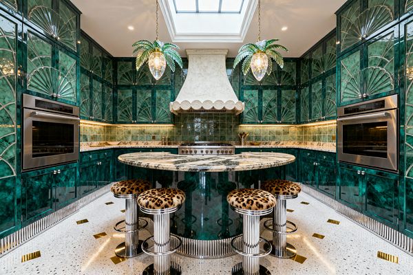 A symmetrical kitchen wrapped in glossy emerald cabinetry with Art Deco fan inlays centers on a range beneath a scalloped white plaster hood, lit from above by a skylight. An oval island with boldly veined marble pairs with chrome fluted stools upholstered in leopard print, terrazzo floors with brass accents, jade square-tile backsplash, warm under-cabinet lighting, stainless wall ovens on both sides, and two palm-shaped glass pendants for Hollywood Regency glamour.