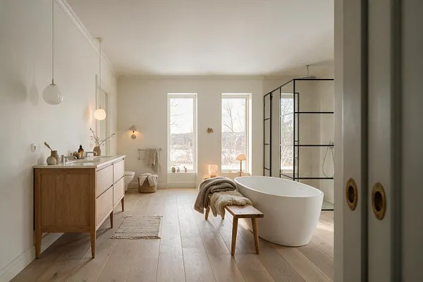 A spacious Scandinavian-style master bathroom with a light wood vanity on the left, a freestanding white oval bathtub in the center, and a black-framed glass shower on the right. Tall narrow windows bring in soft daylight, while neutral walls, pale wood floors, and cozy textiles create a calm, airy atmosphere.