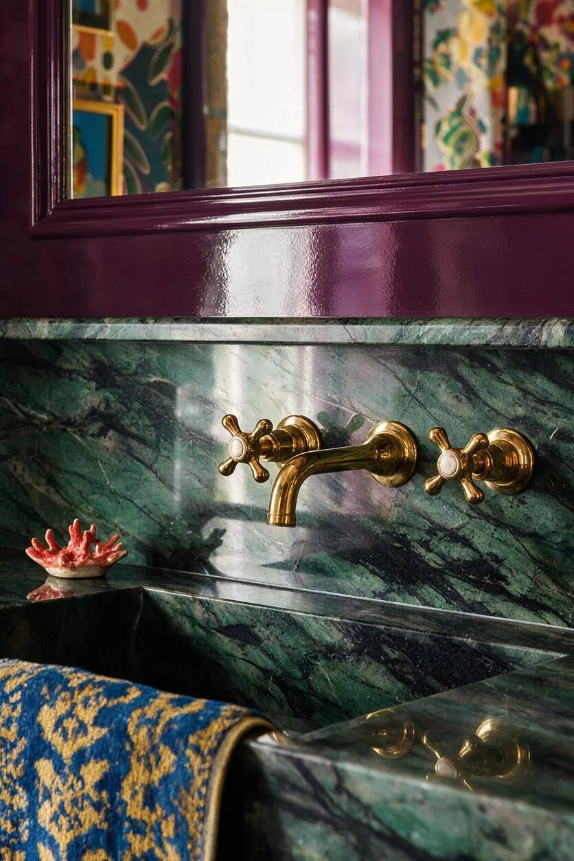 A close-up of a dramatic bathroom sink area with deep green marble or stone, glossy plum trim, and polished brass faucet handles and spout. A patterned blue-and-gold textile hangs over the foreground edge, adding to the richly layered maximalist look.
