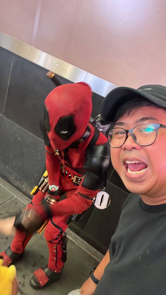A person takes a selfie with a Deadpool cosplayer in a casual setting.