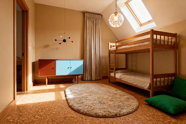A kids’ room with a natural wood bunk bed stands to the right on cork flooring, anchored by an oval cream shag rug. Opposite, a low mid-century cabinet with terracotta and sky-blue sliding doors sits under an atomic mobile; a bubble-glass globe pendant hangs near a skylight in the sloped ceiling, while tan walls, oak trim, a full-length beige linen curtain, and a bright green floor cushion complete the warm, sunlit space.