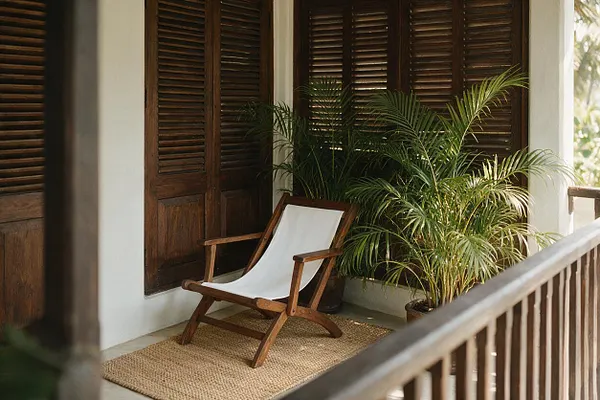 A shaded tropical veranda with dark wooden louvered shutters, a teak folding lounge chair with a white sling seat, potted palm plants, and a woven natural fiber rug on a pale floor. A wooden railing frames the foreground, creating a calm, breezy outdoor retreat.