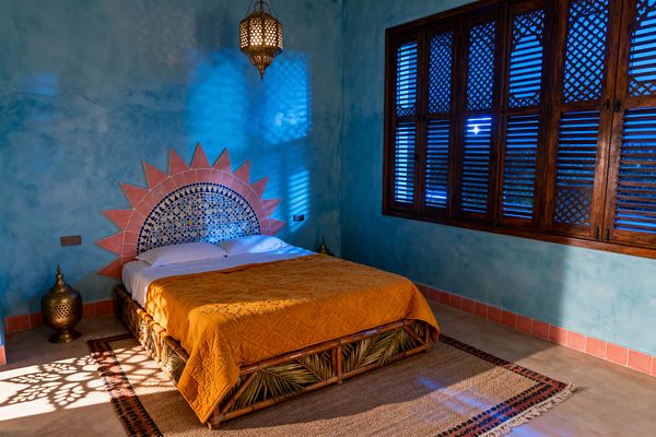 A Moroccan bedroom with turquoise tadelakt walls features a low bamboo bed dressed in a saffron quilt before a half-dome zellige headboard edged with salmon terracotta sun rays. A pierced brass pendant casts patterned light onto a jute rug and cement floor, while carved wooden shutters with lattice panels line one wall and filter deep blue evening light; a small brass floor lantern sits beside the bed.
