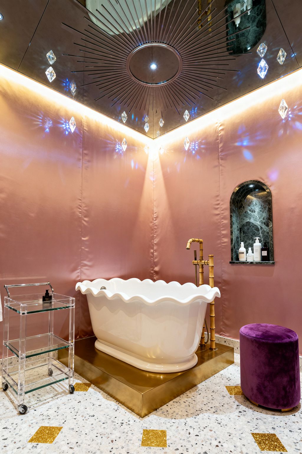 A Hollywood Regency bathroom features a white scalloped freestanding tub on a brushed brass plinth with a bamboo-style brass floor faucet. An arched niche lined in dark green marble holds bottles. The terrazzo floor has gold inlay squares. A clear lucite trolley and a deep purple velvet ottoman sit nearby. Blush pink, satin-sheen walls meet a mirrored ceiling with a sunburst motif, crystal drops, and warm cove lighting that casts prismatic reflections.