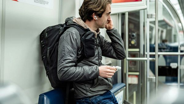 A man is sitting in a subway, wearing a backpack and using wireless earbuds while holding a small item in his hand.