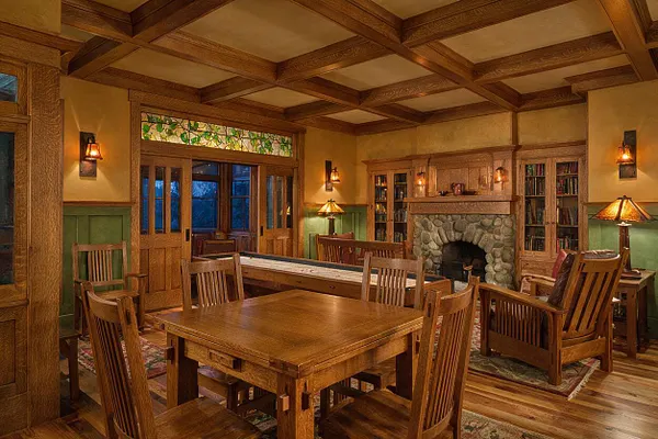 A warm Craftsman-style game room with a coffered wood ceiling, built-in wood cabinetry, and a large stone fireplace centered on the back wall. The room includes a wooden game table, slatted wood chairs, a shuffleboard table, and amber wall sconces, with green wainscoting and stained-glass transom windows adding detail.