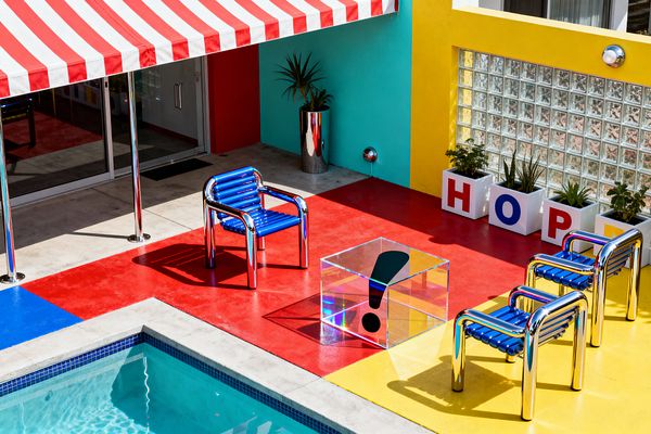 A poolside patio is color-blocked in glossy red, yellow, and blue painted concrete, furnished with chrome-tube chairs that have cobalt slat seats around a clear acrylic cube table marked with a black exclamation point. A red-and-white striped awning shades aluminum sliding doors; adjacent turquoise and canary walls feature round sconces, a glass-block window, and white planters spelling HOPE beside a stainless planter with a small palm.