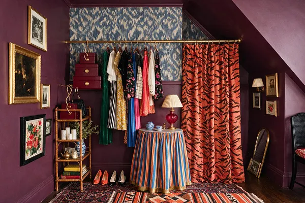 A dramatic closet corner with deep plum walls, a blue patterned accent wall, and a brass clothing rod holding colorful garments. The space includes tiger-print orange curtains, framed artwork, a striped skirted table with a red lamp, and layered rugs, creating a richly eclectic maximalist look.