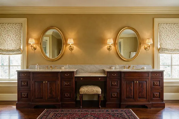 A traditional master bathroom with two dark wood vanities flanking a central makeup seating area, topped with light marble countertops and paired gold-framed oval mirrors. Soft gold walls, wall sconces, floral Roman shades, and warm wood flooring create an elegant, classic atmosphere.