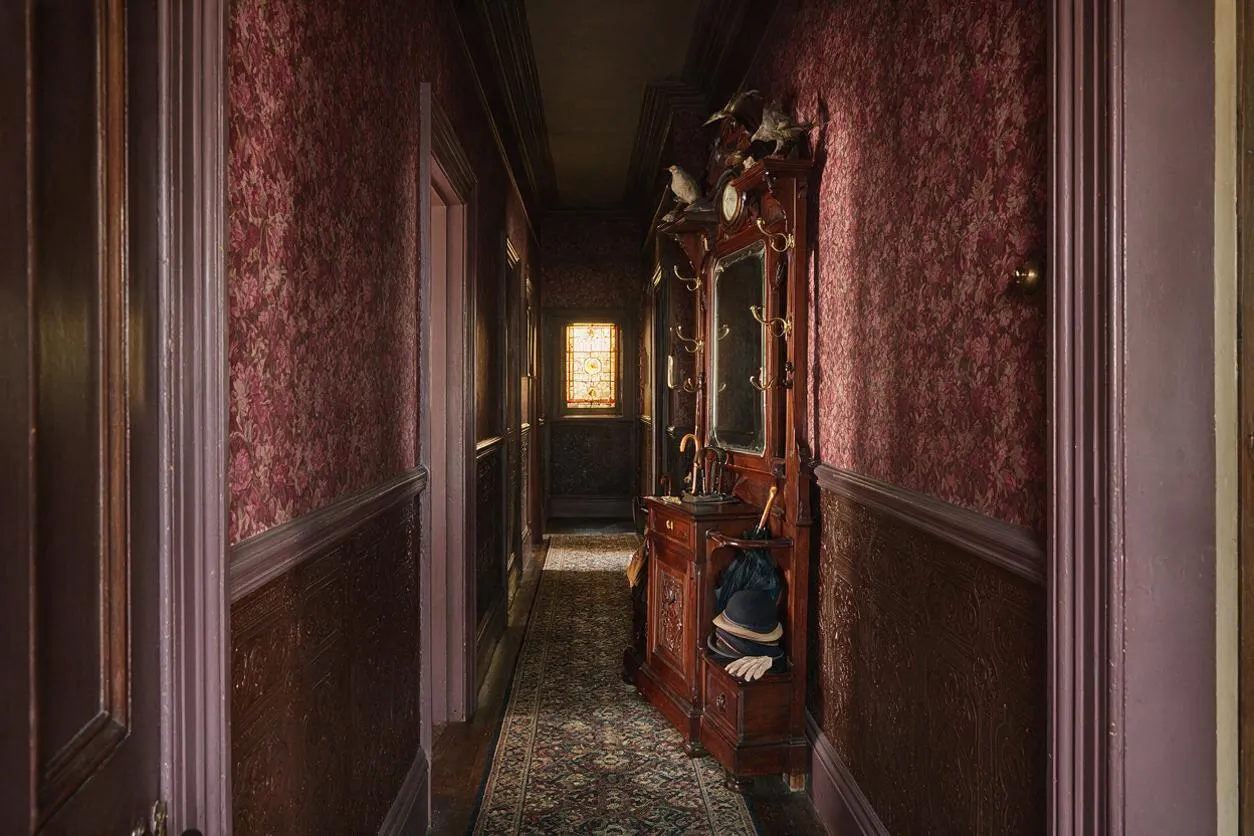 A narrow Victorian entryway with burgundy floral wallpaper, dark wood wainscoting, and a patterned runner rug leading toward a stained-glass window. An ornate carved wood hall stand with a mirror, hooks, and hats creates a formal, antique look.