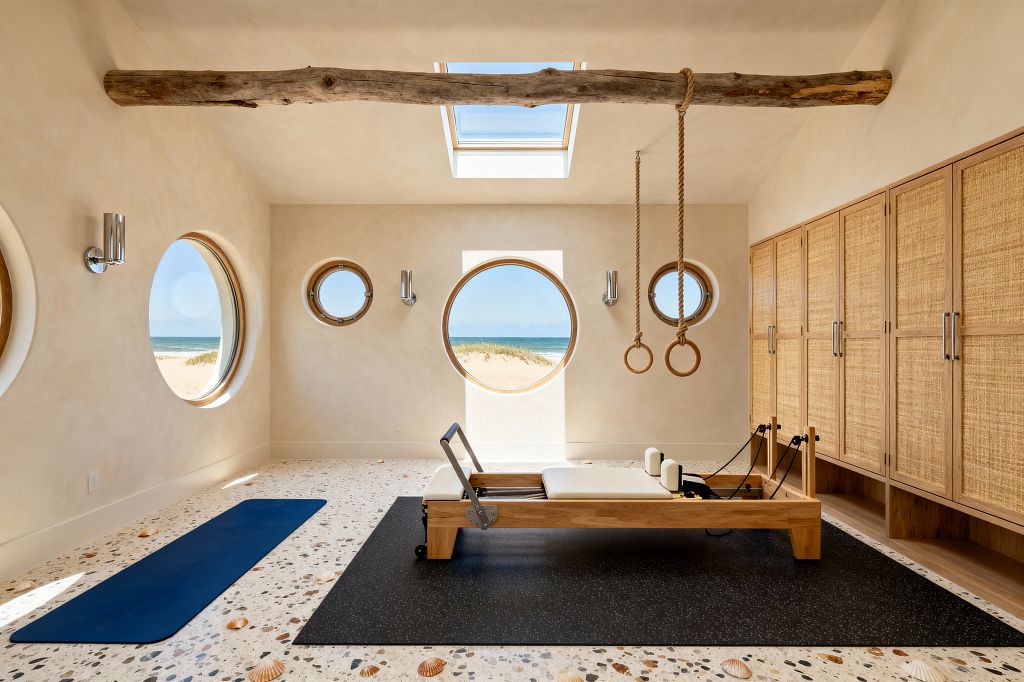 A serene coastal home gym with sandy limewashed walls shows a wood Pilates reformer on a black rubber mat and a blue yoga mat on terrazzo flooring inlaid with seashells. Round porthole windows and a larger central circular window reveal dunes and ocean; a skylight above and chrome sconces provide additional light. A driftwood ceiling beam suspends rope gymnastic rings, and a wall of oak cabinets with woven rattan doors lines the right side.