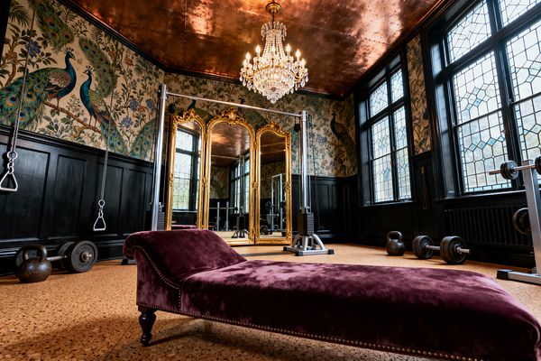A luxurious Victorian-style home gym featuring a deep-plum velvet chaise lounge in the foreground, an ornate gold trifold mirror behind a cable machine, and a crystal chandelier hanging from a burnished copper ceiling. Black wainscoting and peacock-patterned wallpaper line the walls, while leaded-glass windows illuminate cork flooring scattered with kettlebells, barbells, and weight plates.