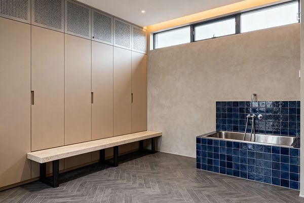 Modern mudroom with a wall of full-height matte taupe cabinets topped by perforated metal panels and a long light stone bench on black steel legs. A gray herringbone tile floor leads to a stainless utility basin framed in glossy navy square tiles with a wall-mounted faucet and hand spray. Smooth plaster walls, a slim clerestory window, and warm LED cove lighting create a clean, functional space.