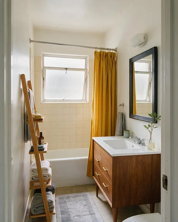 A narrow master bathroom with a walnut vanity, white countertop, black-framed mirror, and mustard-yellow shower curtain. A wooden ladder shelf holds towels and bath products beside a small tiled tub and frosted window, creating a warm mid-century modern look.