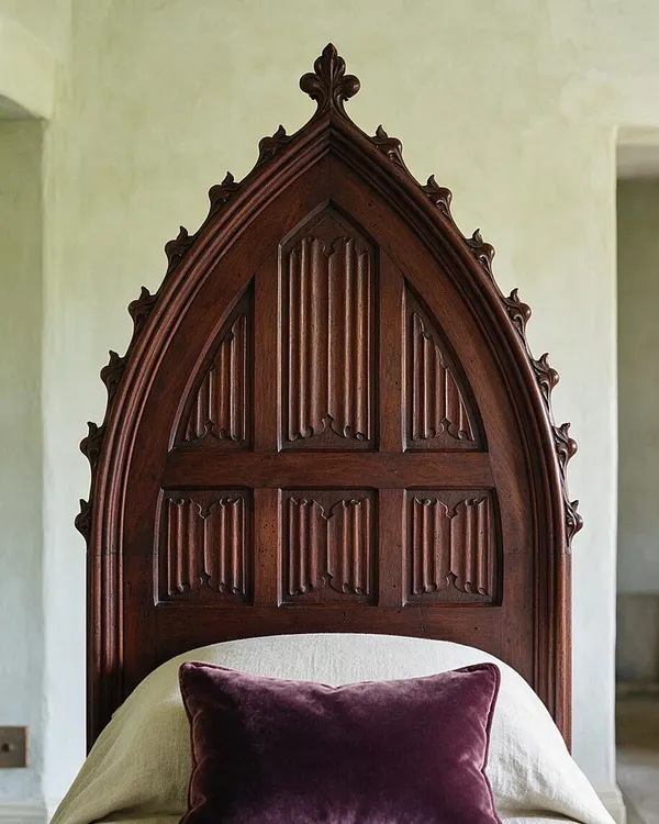 A bed with a tall, pointed Gothic-style carved wooden headboard in dark stained wood. The headboard features arched paneling and ornate finial-like detailing, set against pale walls with neutral bedding and a deep plum velvet pillow.