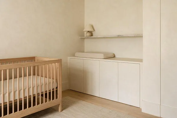 A serene minimalist nursery with a light wood crib on the left, white bedding, and a long built-in storage cabinet along the back wall. A simple shelf with a small lamp sits above the cabinet, while soft beige walls, pale wood flooring, and a neutral rug create a calm, uncluttered atmosphere.