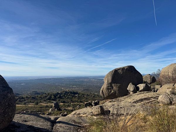 Rocky hilltop with large boulders overlooking a densely forested landscape under a blue sky with wispy clouds and contrails.