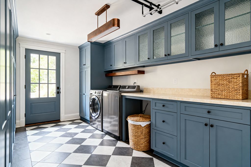 Transitional laundry room featuring dusty blue Shaker cabinets with textured-glass uppers, matte black hardware, and a long light beige stone countertop; a stainless washer and dryer sit beneath a walnut floating shelf. The room is grounded by a diagonal charcoal-and-white checkerboard tile floor and brightened by a multi-lite blue exterior door, white walls, and a slim bronze linear pendant overhead.
