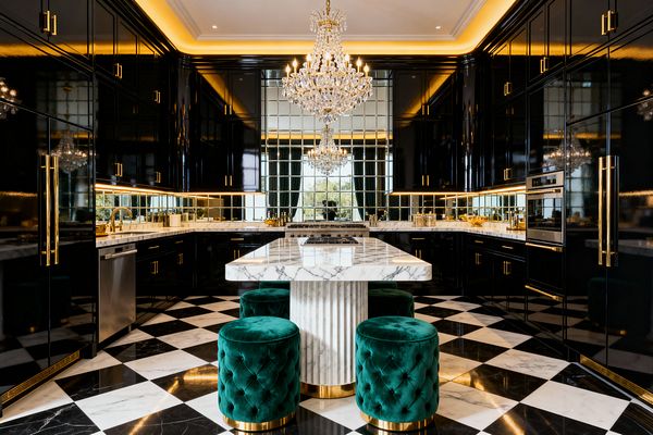 A glamorous kitchen with high-gloss black lacquer cabinets accented by brass pulls, white marble counters, and a marble island with a fluted base. Emerald tufted velvet stools with gold bases sit on a black-and-white checkerboard marble floor, while mirrored backsplash tiles, cove and under-cabinet lighting, and two crystal chandeliers create sparkle; stainless appliances and a window at the end complete the symmetrical layout.