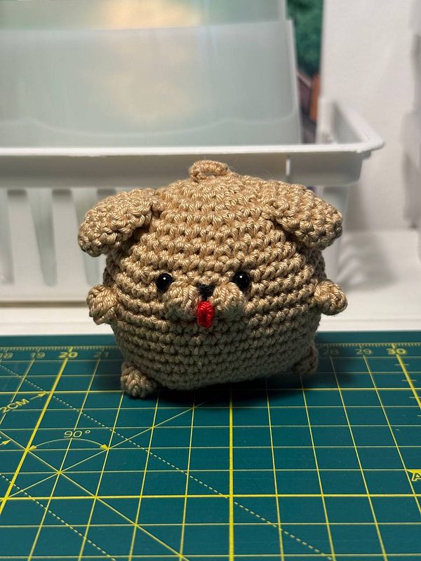 A crocheted critter resembling a small, round animal with a cute face is displayed on a cutting mat.