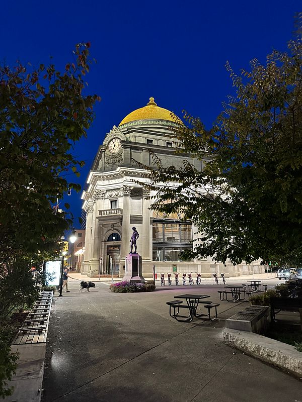 A historic building with a golden dome is illuminated at night, surrounded by trees and outdoor seating.