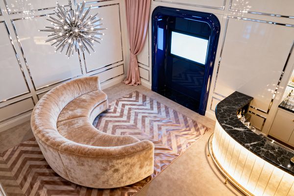 A glamorous media room features a sweeping curved velvet sectional in champagne beige atop a chevron, satin-sheen rug. A polished-chrome starburst chandelier hangs above cream paneled walls with mirror-bright trim, while a curved, backlit bar with a black marble countertop and brass details faces a screen set within a high-gloss navy lacquer niche beside blush-pink drapery.