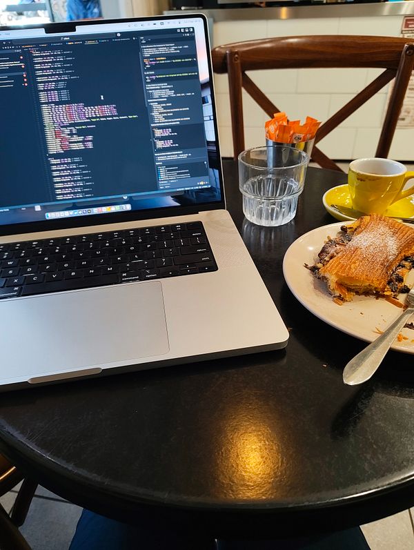 A laptop displaying code is set on a café table alongside a pastry and a cup of coffee.