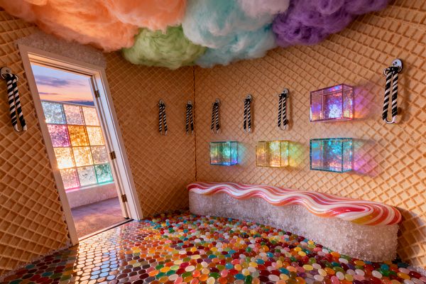 A whimsical mudroom styled like a candy shop. Tan waffle-cone patterned walls hold black-and-white striped hooks. A long bench shaped like ribbon candy rests on a sparkling, sugar-like base. The glossy floor is a mosaic of multicolored jellybean shapes. The ceiling is filled with fluffy pastel clouds in peach, mint, aqua, and violet. Iridescent wall boxes glow with rainbow light, and a doorway opens to a space with colorful glass-block windows.