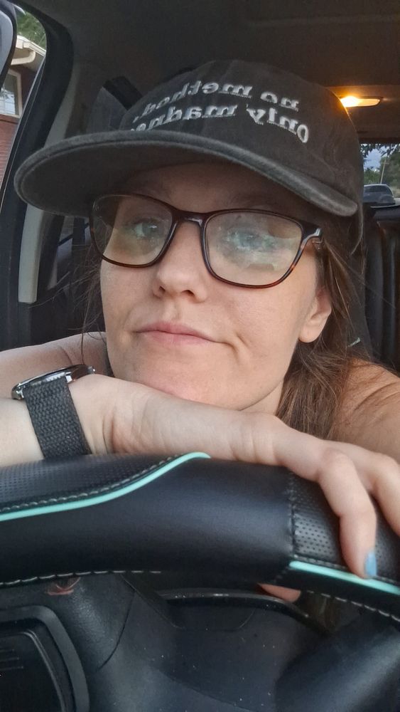 A tired individual rests their chin on their hand while sitting in a car, wearing a cap and glasses.