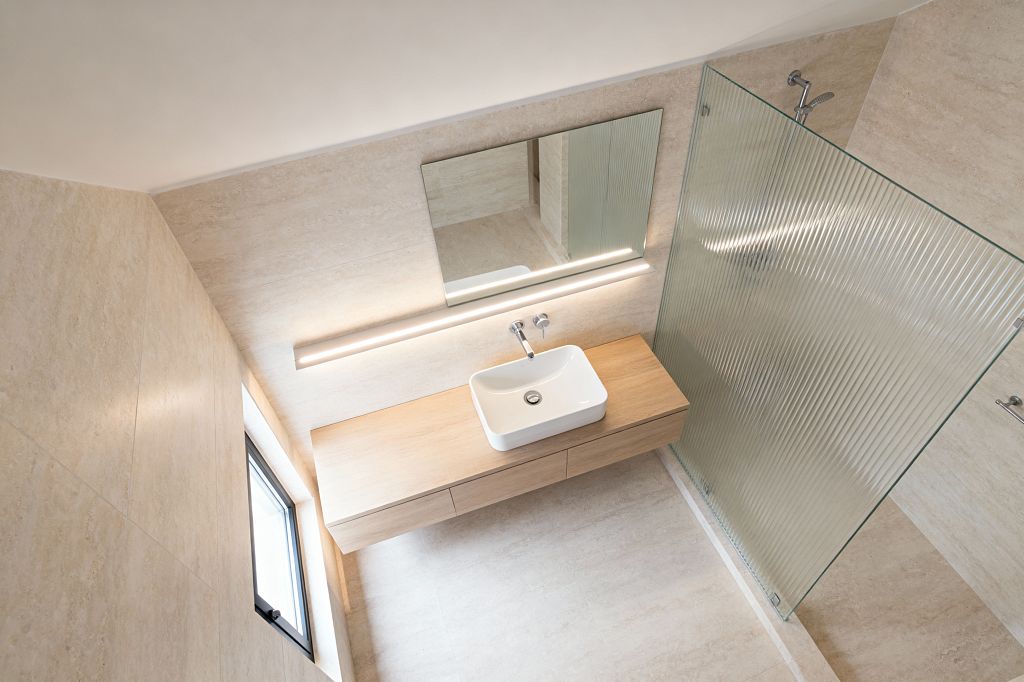 A minimalist bathroom clad in warm beige stone-look tiles features a floating light-wood vanity with a white rectangular vessel sink and wall-mounted chrome faucet below a wide frameless mirror. A slim linear LED runs along the wall, and a ribbed glass shower screen defines the walk-in area with a simple chrome shower set. A small black-framed window brings daylight, and integrated drawers keep the space uncluttered.
