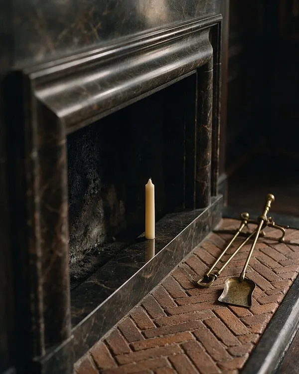 A dark, moody fireplace in black marble with a polished mantel and recessed firebox, styled in a Dark Academia aesthetic. A single cream taper candle stands on the hearth, and brass fireplace tools rest on a red herringbone brick floor, adding warm metallic contrast to the charcoal interior.