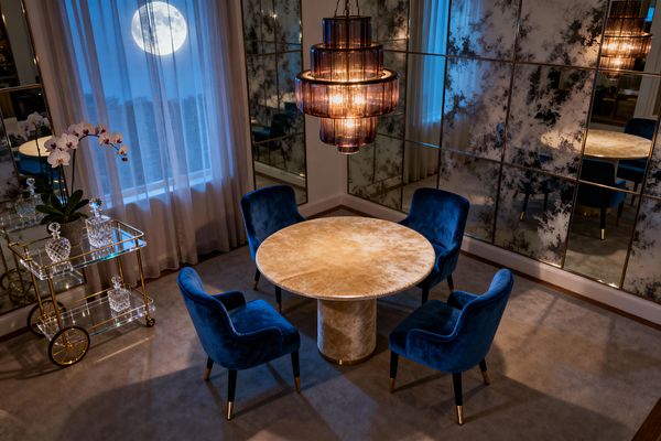 A round dining table with a champagne-lacquer finish sits under a tiered smoked-glass chandelier, surrounded by four sapphire-blue velvet chairs with brass-capped legs. One wall is clad in antiqued mirror panels, a brass-and-glass bar cart holds crystal decanters beside an orchid, and plush taupe carpeting and sheer ivory curtains, with a moonlit view beyond, create a warm yet moody atmosphere.