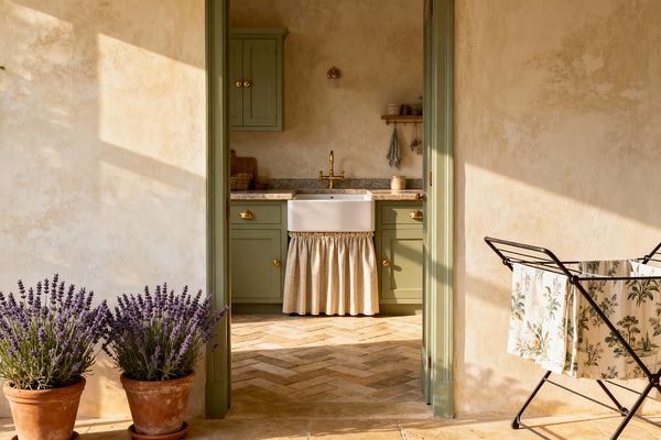 A sunlit French Country laundry room viewed through a doorway shows sage-green Shaker cabinets with brass knobs and pulls, a white fireclay farmhouse sink with a beige linen skirt, and a speckled stone countertop topped by a brass bridge faucet. Warm limewashed plaster walls, a herringbone terracotta floor, a small brass sconce, an open wooden shelf with jars, potted lavender, and a floral fabric laundry hamper complete the rustic scene.