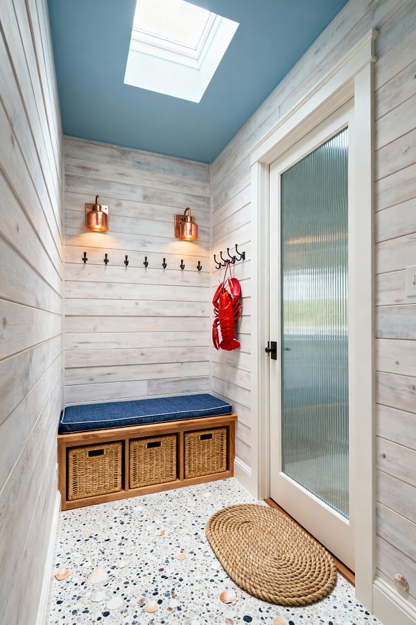 A compact coastal mudroom clad in whitewashed shiplap features a sky-blue ceiling with a large skylight that floods the space with daylight. A built-in wood bench with a navy cushion sits over three rattan baskets, with a row of matte black hooks and two copper-and-glass sconces above; a fluted-glass door with black hardware opens beside a rope doormat, while speckled terrazzo flooring with seashell inlays and a red lobster decoration complete the nautical look.