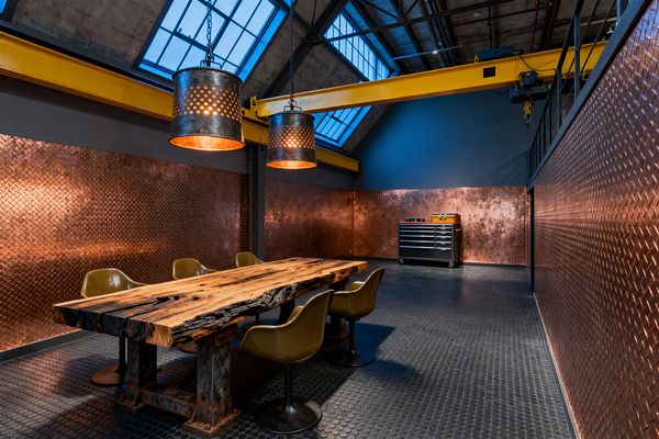 Industrial dining space in a tall, skylit loft with exposed steel trusses and a bright yellow overhead gantry crane. A live-edge wood slab table on heavy steel trestles is surrounded by olive swivel chairs. Copper diamond-plate panels clad the walls, reflecting warm light from two perforated drum pendants with copper interiors. The floor is black rubber coin tile, and a rolling tool chest stands along the back wall, reinforcing the workshop character.