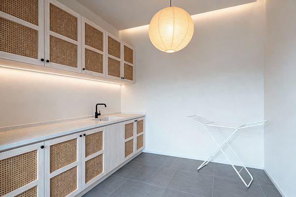 A minimalist Scandinavian laundry room with light wood cabinets featuring cane webbing panels runs along the left wall, topped by a pale quartz countertop and an integrated sink with a matte black faucet. Warm under-cabinet strip lighting and a ceiling cove glow illuminate white walls, while a round paper lantern hangs overhead; large gray floor tiles and a white folding drying rack keep the space calm and uncluttered.