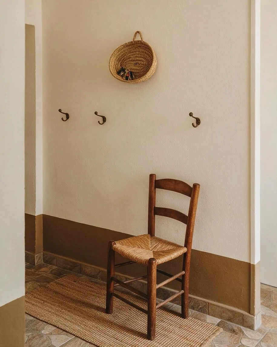 A narrow Mediterranean-style entryway with cream stucco walls, a brown painted lower wall band, and a patterned stone tile floor. A simple wooden chair with a woven rush seat sits on a natural jute rug, with three wall hooks and a hanging woven basket above it.