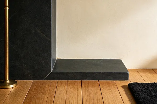 A Mid-Century Modern room detail featuring a dark stone fireplace base with a smooth, angular profile, set against a cream wall and warm oak plank flooring. A slim brass lamp stand appears at the left edge, and a black textured rug anchors the lower right corner.