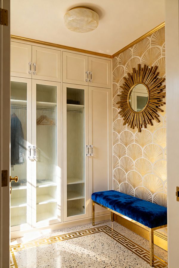 A glamorous mudroom features tall cream cabinetry with reeded-glass doors and clear acrylic pulls, while gold fan-pattern wallpaper holds a gilded sunburst mirror above a royal blue velvet bench with a brass base. A terrazzo floor framed by a gold Greek key inlay, an alabaster flush-mount light, and gold crown molding add warm illumination, with open cubbies and a hanging rail visible inside the cabinets.