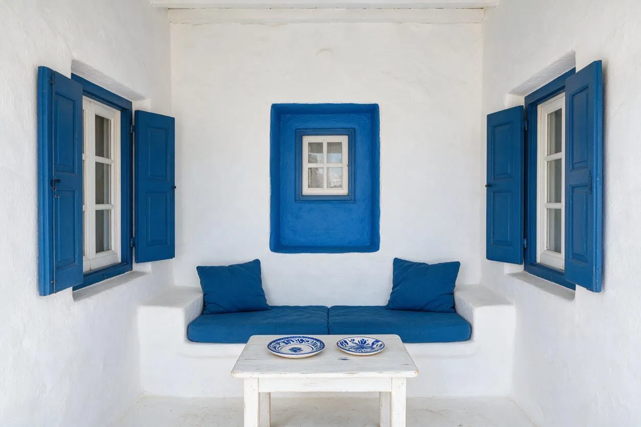 A compact Mediterranean-style sunroom with white plaster walls, cobalt blue window shutters, and a built-in bench topped with blue cushions and matching pillows. A small white table with two patterned ceramic plates sits in front, creating a bright, simple coastal seating nook.